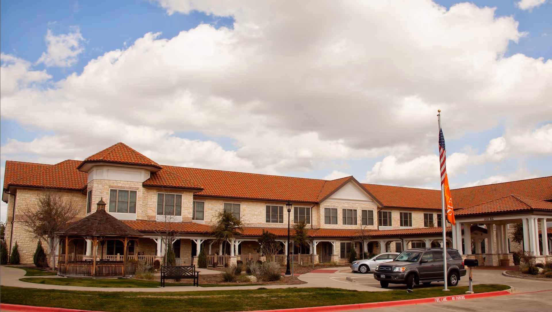 Exterior front view of a two-story senior living facility with a red tile roof, gazebo, flagpole and parked cars.