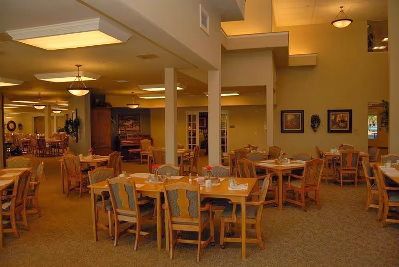 A spacious dining room in a senior living facility with multiple wooden tables and chairs arranged neatly. Each table is set with napkins, utensils, and small flower vases. The room has warm lighting with ceiling fixtures and is decorated with framed pictures and plants. There are columns and a piano visible in the background.