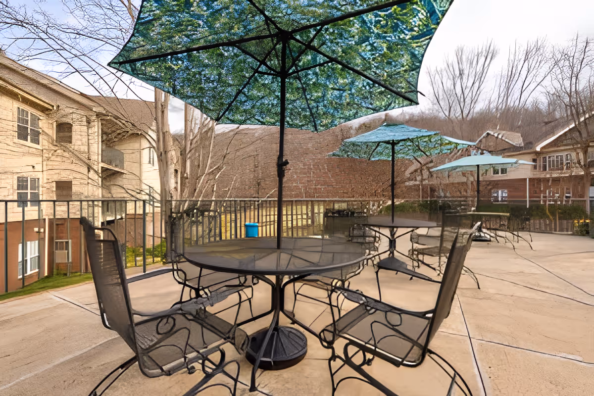 Outdoor patio area with several black metal tables and chairs, each table shaded by a large umbrella with a leafy tree pattern. Surrounding the patio are buildings and leafless trees in the background.