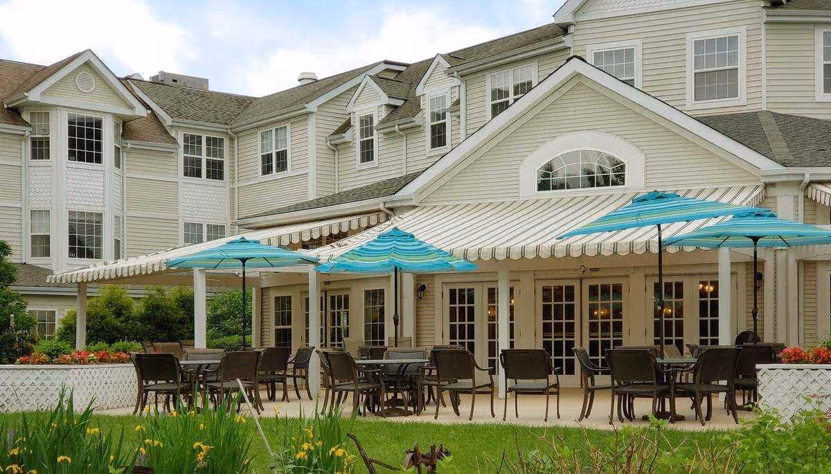 Outdoor patio area of a senior living facility with multiple tables and chairs under blue umbrellas. The building behind has large windows and a striped awning covering the patio. There is greenery and flowers in the foreground.