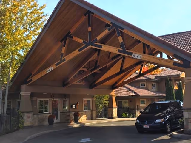 Covered porte-cochere entrance with exposed timber beams in front of a senior living building and a parked car.