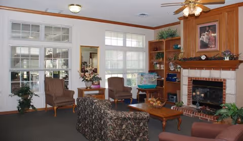 Cozy senior living lounge with a floral sofa, armchairs, coffee table, built-in shelves and a fireplace beneath a ceiling fan.