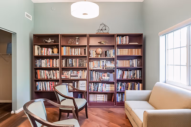 Sunlit reading room with tall bookshelves, two chairs and a sofa beside a window.