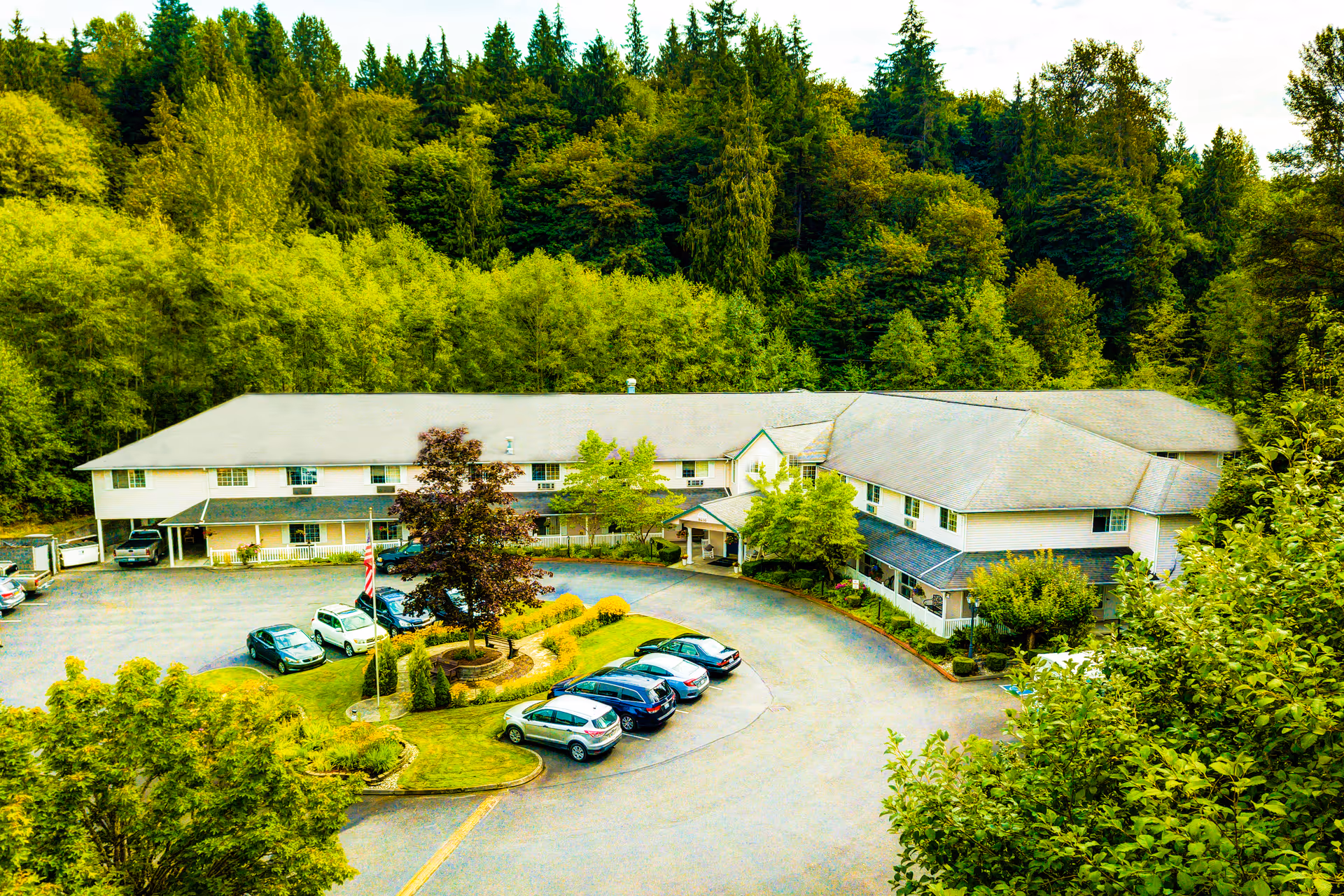 Aerial view of Cascade Valley Senior Living facility surrounded by dense green trees. The building is two stories with a gray roof and light-colored exterior walls. There is a circular driveway with several parked cars and a landscaped area with a tree and shrubs in the center.