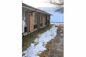 Exterior side view of a one-story brick building with windows along a sidewalk and patches of melting snow on the grass.