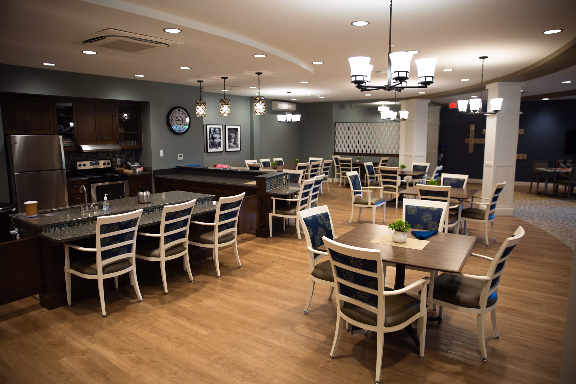 A spacious dining area in an assisted living facility featuring multiple tables with chairs arranged around them. The room has wooden flooring, modern pendant and chandelier lighting, and a kitchen area with stainless steel appliances including a refrigerator and stove. The walls are decorated with framed pictures and a clock.