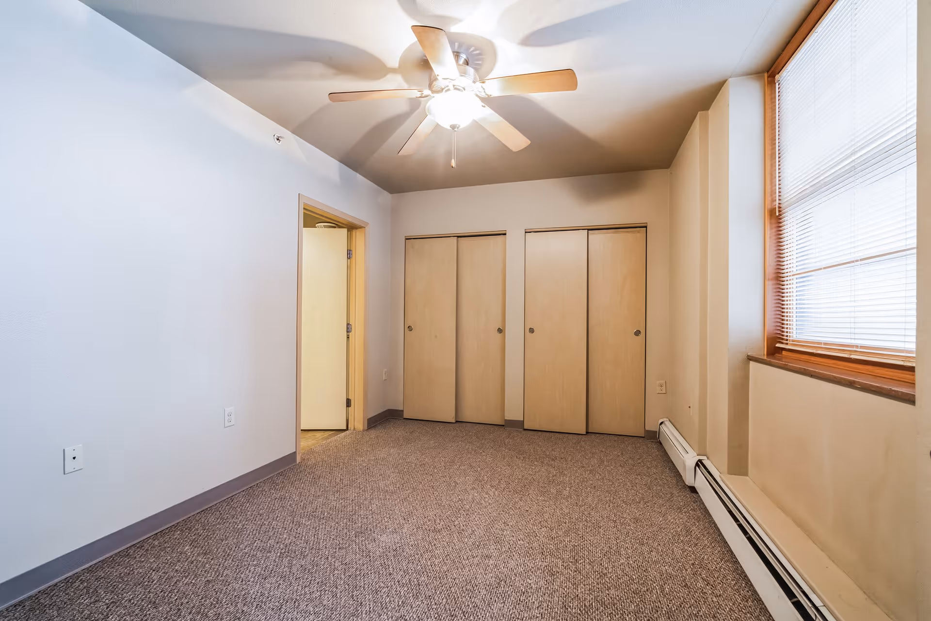 Empty room with beige carpet, two closed wooden closets, a ceiling fan with light, a window with blinds, and an open door leading to another room.