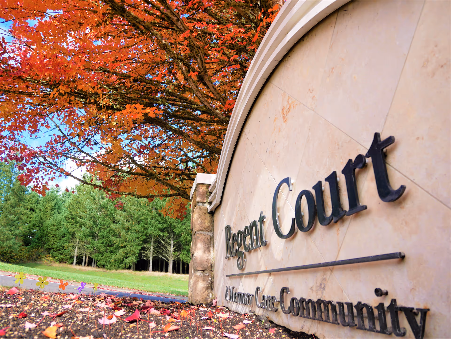 Stone sign for Regent Court Assisted Living Care Community with autumn trees featuring bright orange and red leaves in the background and fallen leaves on the ground.