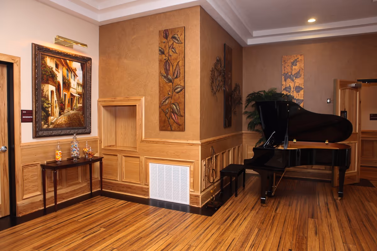 An elegant interior corner of a senior living facility featuring a polished black grand piano with a matching bench, wooden paneled walls with decorative leaf artwork, a small table with jars of candy, a framed painting of a quaint street scene, and warm wooden flooring.