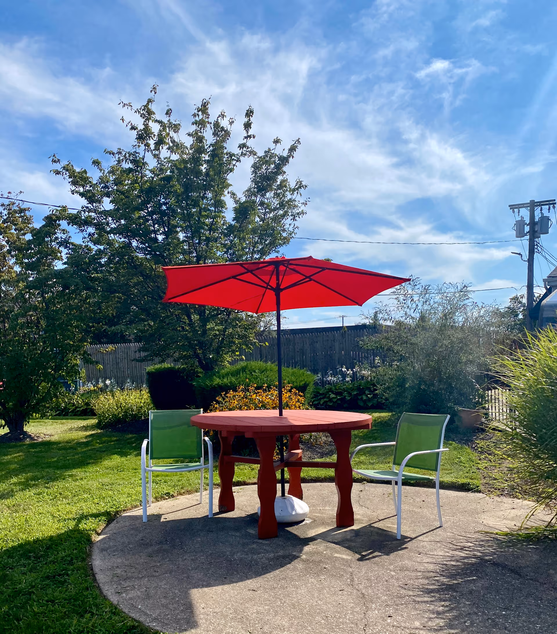 Outdoor seating area with a round red table and a red umbrella, accompanied by two green chairs on a concrete patio surrounded by grass, trees, and shrubs under a partly cloudy blue sky.