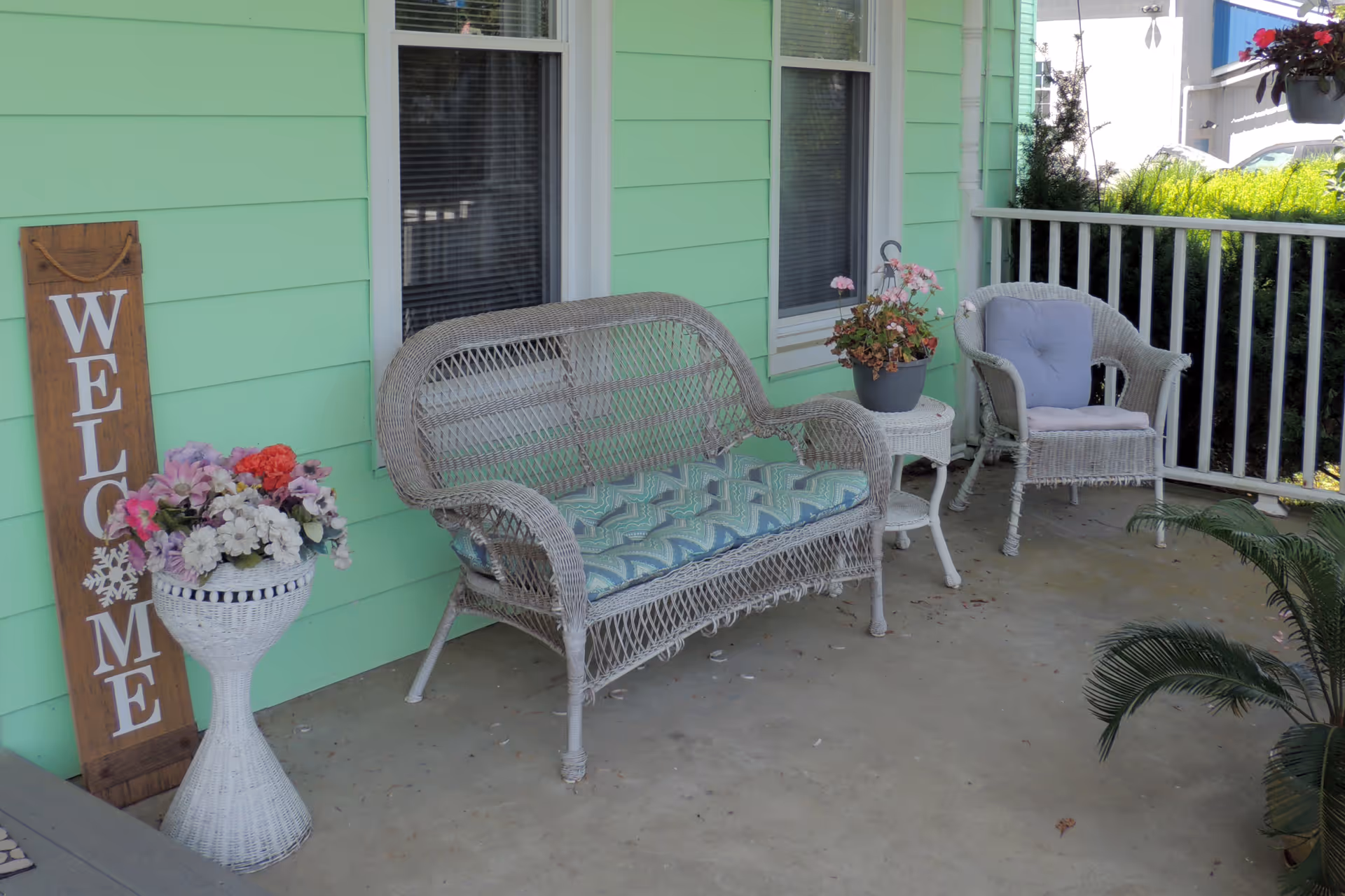 A porch area with light green siding, featuring a white wicker loveseat with a patterned cushion, a matching wicker chair with a blue cushion, a small white side table with a potted plant, a white wicker flower stand with a bouquet of colorful flowers, and a wooden welcome sign leaning against the wall.