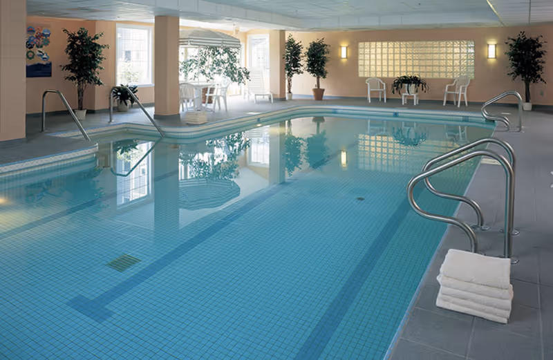 Indoor swimming pool with clear blue water, metal handrails, and a stack of white towels on the poolside. The room has large windows letting in natural light, potted plants, white chairs, and a table with an umbrella near the windows.