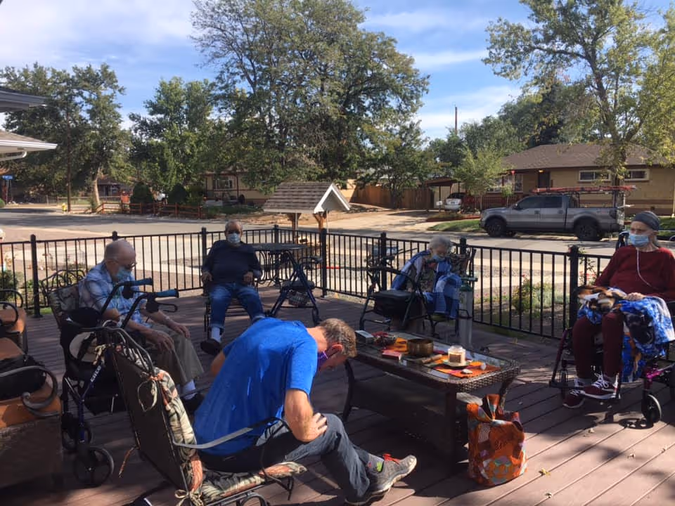 A group of elderly individuals sitting outdoors on a wooden deck, some in wheelchairs and others in chairs, all wearing face masks. They are gathered around a low table with various items on it. Trees, houses, and parked vehicles are visible in the background under a partly cloudy sky.