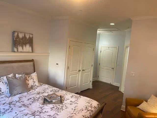 A bedroom with a bed featuring a floral patterned comforter and multiple pillows. There is a small tray on the bed. The room has light-colored walls, a piece of abstract artwork above the bed, wooden flooring, and several closed white doors. A yellow armchair with a cushion is partially visible on the right side.