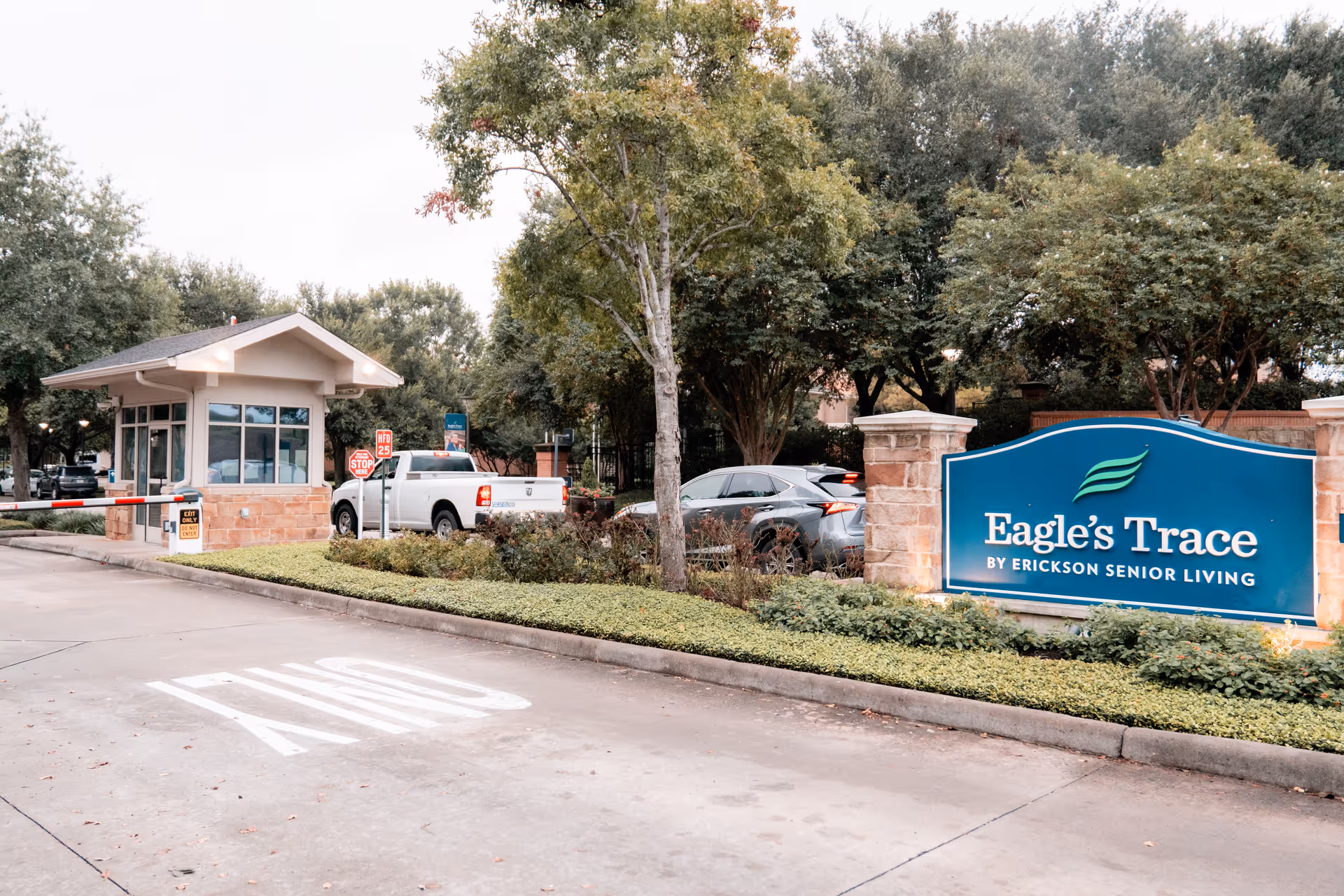 Entrance to Eagle's Trace senior living facility showing a guardhouse, a stop sign, a white pickup truck, a gray SUV, and a large blue sign with the facility name surrounded by greenery and trees.