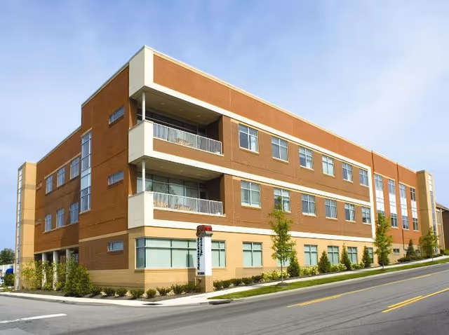 Exterior view of a three-story brick and beige building with multiple windows and balconies, situated on a street corner with landscaping and a clear sky above.