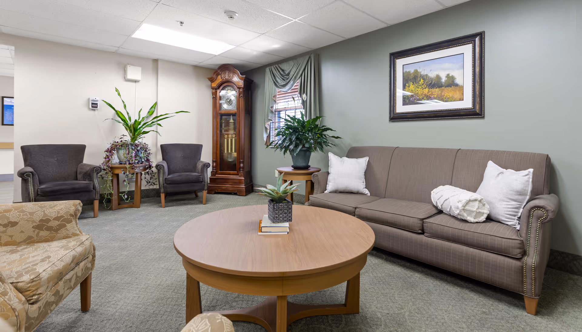 A cozy sitting area in a senior living facility with a brown sofa adorned with white pillows, two dark armchairs, a patterned beige armchair, a round wooden coffee table with a small plant and books, a tall wooden grandfather clock, and green plants near a window with drapes. A framed landscape painting hangs on the light green wall.