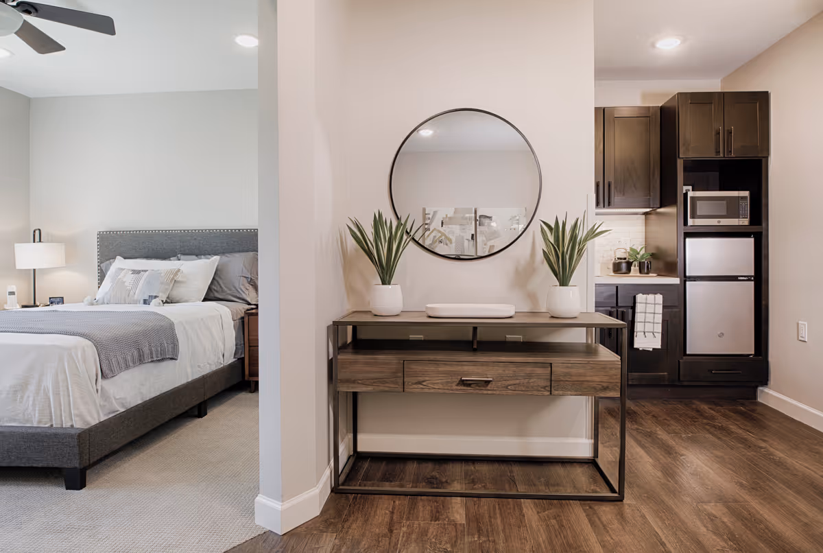 View of a modern assisted living unit showing a bedroom with a gray upholstered bed, white and gray bedding, a nightstand with a lamp, and a ceiling fan. Adjacent to the bedroom is a small kitchen area with dark wood cabinets, a microwave, and a mini refrigerator. In the foreground, there is a wooden console table with a metal frame, decorated with two potted plants and a round mirror mounted on the wall above it. The flooring is a combination of carpet in the bedroom and wood in the kitchen and hallway.