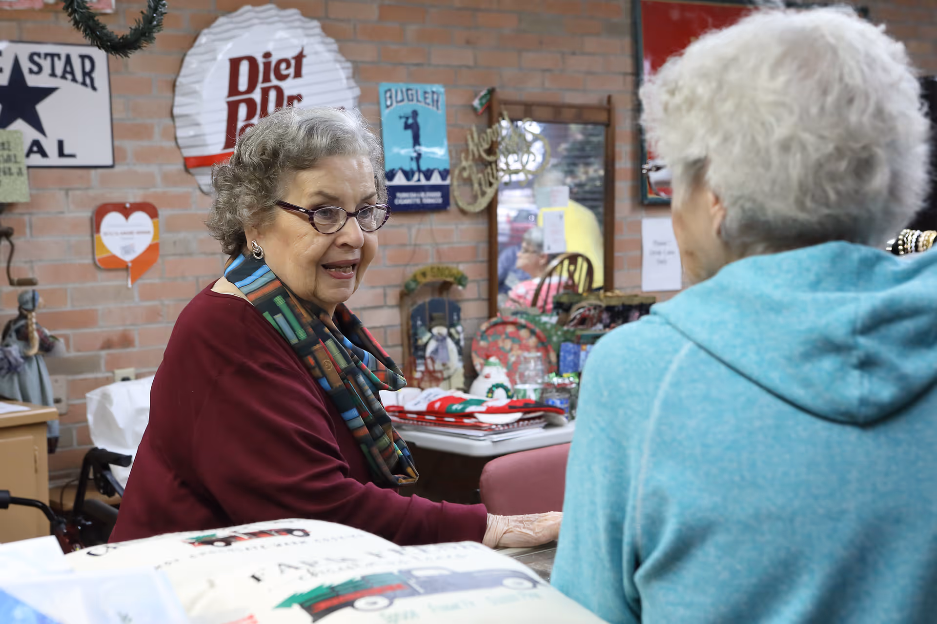 Two elderly women engaged in conversation in a cozy room decorated with vintage signs and holiday decorations. One woman, wearing glasses and a colorful scarf, is facing the camera, while the other woman, with white hair and a blue hoodie, has her back to the camera. The room has a brick wall and various items on a table, suggesting a communal or activity area.