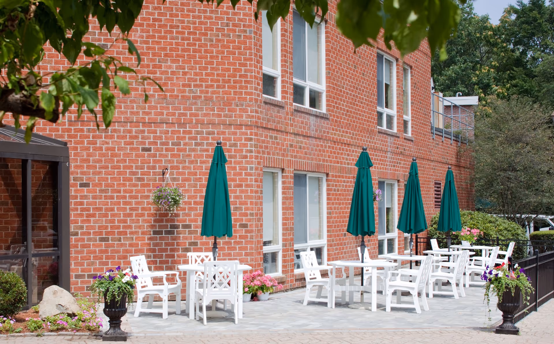 Outdoor patio area with white tables and chairs, each table shaded by a closed green umbrella, set against a red brick building with several windows. There are hanging flower baskets and potted plants decorating the space, with trees and greenery in the background.