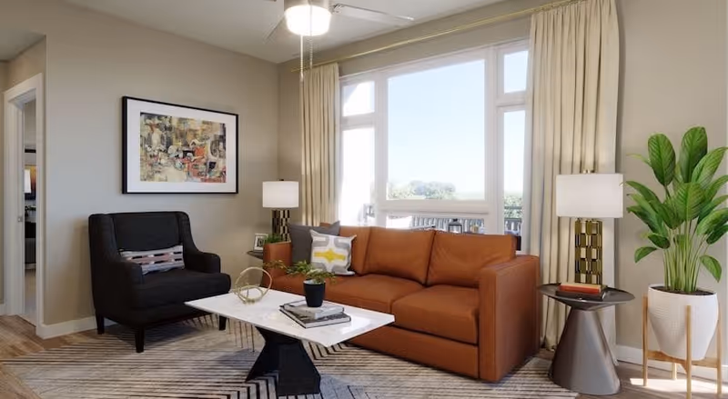 A bright living room with a large window covered by beige curtains. The room features a brown leather sofa with decorative pillows, a black armchair with a patterned cushion, a white marble coffee table with books and a small plant, two modern side tables with lamps, a large green potted plant, and a framed abstract painting on the wall.