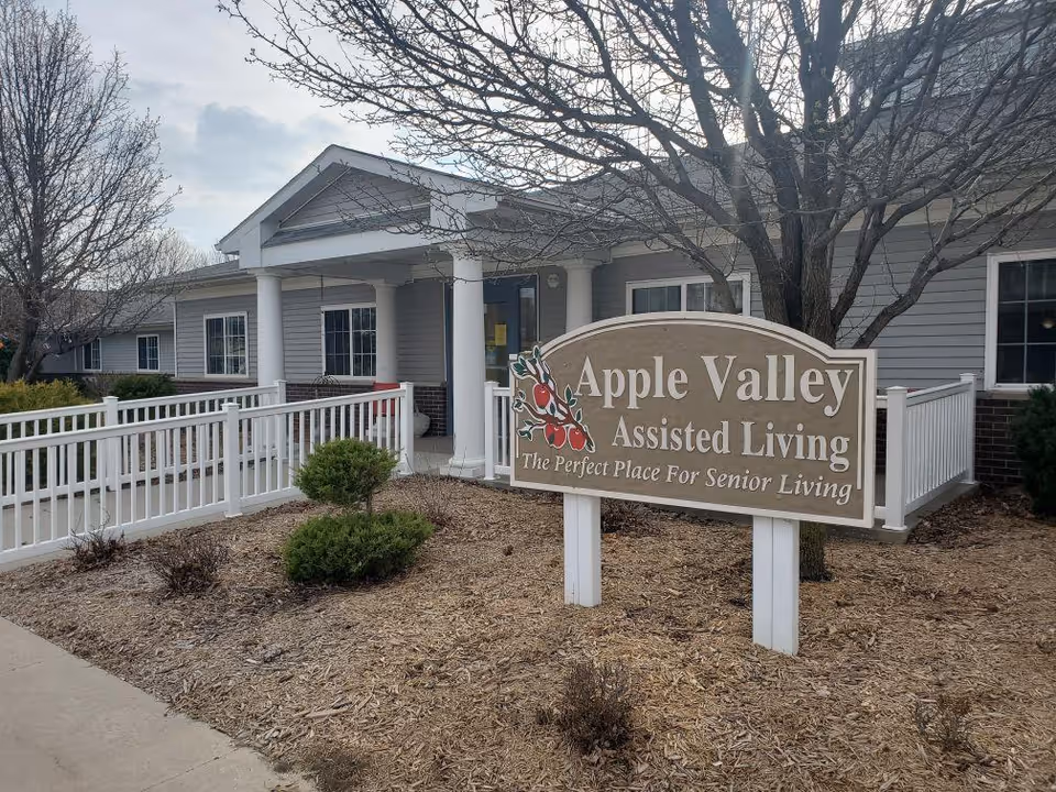 Exterior view of Apple Valley Assisted Living facility showing the entrance with white columns and a white railing ramp. In front, there is a large sign that reads 'Apple Valley Assisted Living The Perfect Place For Senior Living' with an illustration of red apples. Leafless trees and some small bushes are visible around the building.