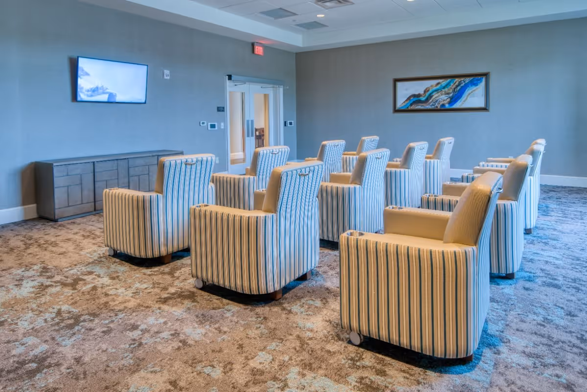 A room with multiple striped armchairs arranged in rows facing a wall-mounted television. The room has a carpeted floor, a sideboard under the TV, a framed abstract painting on the wall, and double glass doors at the back.