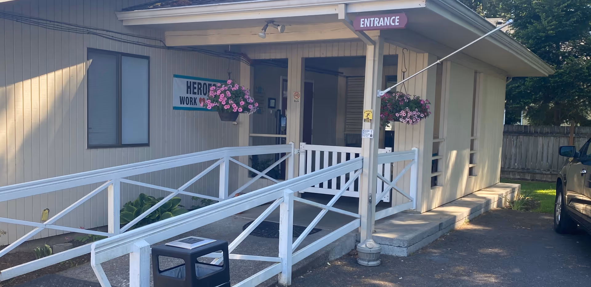 Exterior entrance of a care facility showing a covered doorway with an 'ENTRANCE' sign, a wheelchair ramp, and hanging flower baskets.
