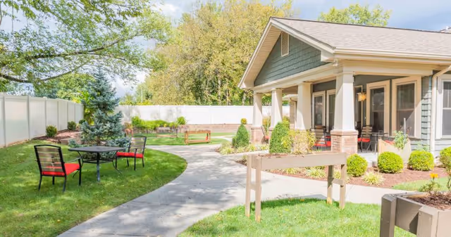 Sunny courtyard with a paved walkway, lawn seating with red‑cushioned chairs, landscaping, and a covered porch of a light-gray building.