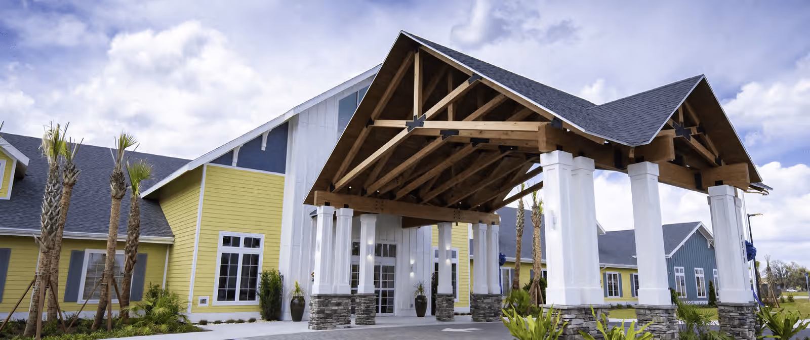Exterior view of Inspiritás of Wildwood facility showing a large covered entrance with wooden beams supported by white columns with stone bases, surrounded by palm trees and landscaped greenery under a partly cloudy sky.
