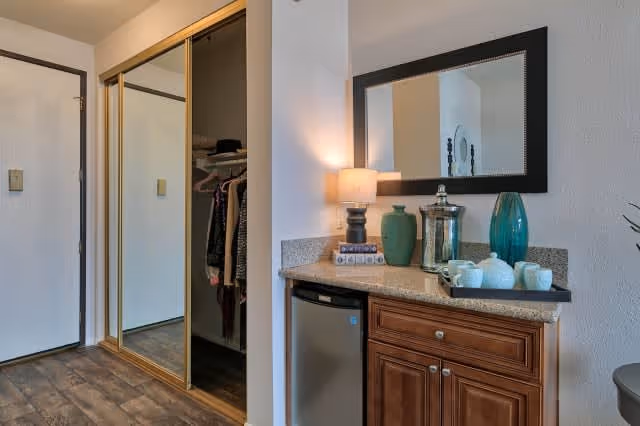 Interior view of a room featuring a wooden cabinet with a granite countertop holding decorative items including a lamp, vases, and a tea set. Above the cabinet is a large rectangular mirror. To the left, there is a closet with sliding mirrored doors reflecting part of the room. The floor is wooden, and the walls are painted white.