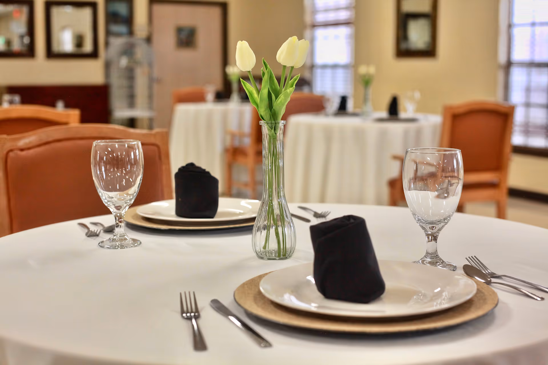 A neatly set dining table with plates, wine glasses, black napkins and a vase of white tulips in a dining room.
