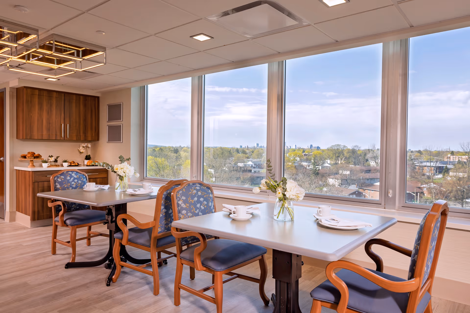 A bright dining area with large windows offering a scenic view of trees and buildings outside. The room features two tables with blue cushioned chairs, each table set with white cups, plates, and napkins. A vase with white flowers is placed on each table. In the background, there is a wooden cabinet with pastries and decorative items on the countertop. The ceiling has modern rectangular light fixtures.