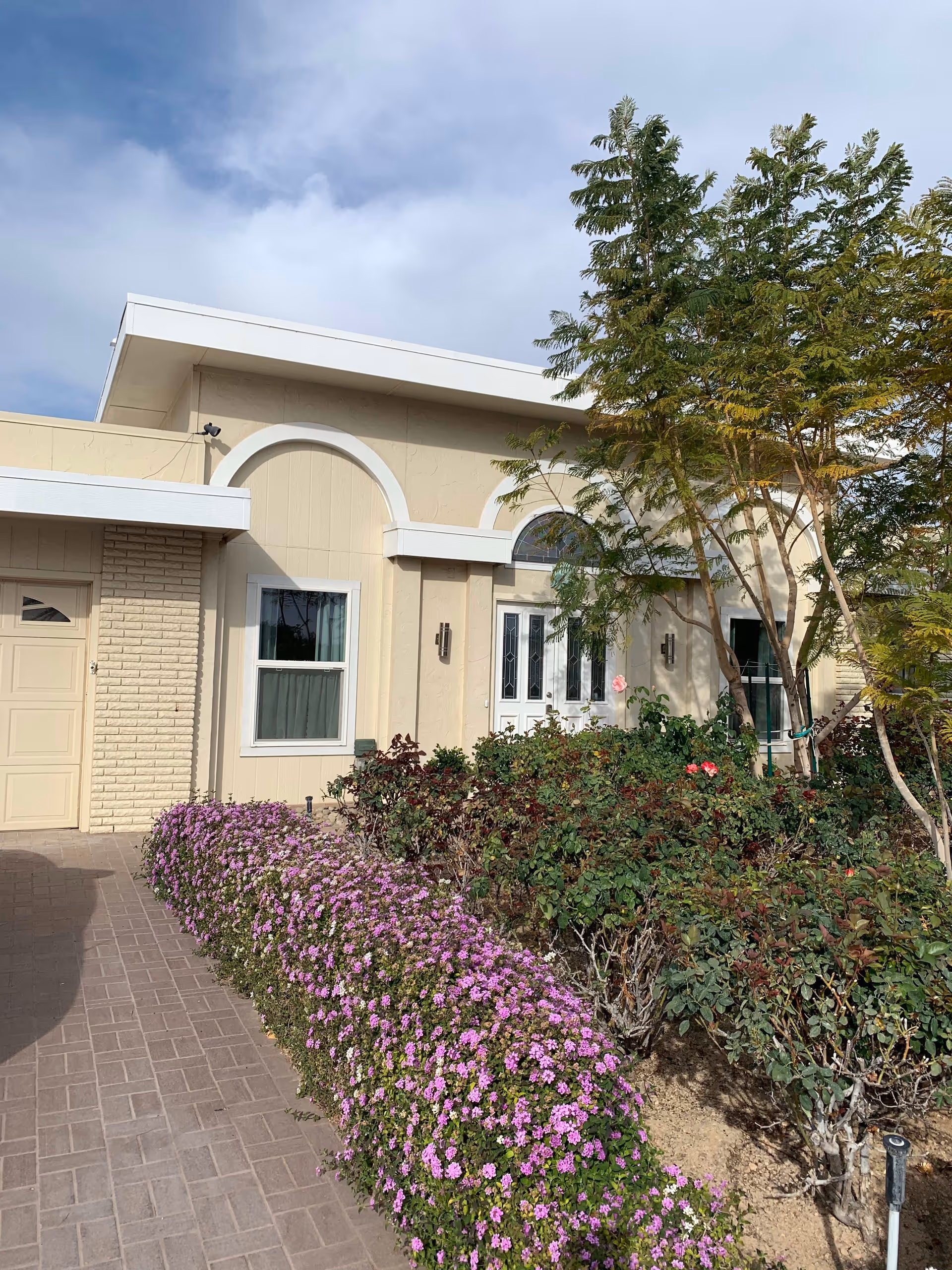 Exterior view of a beige assisted living facility building with white trim, featuring a white double door entrance and a window. In front of the building, there is a garden with green shrubs, pink flowers, and a tree. The sky is partly cloudy.