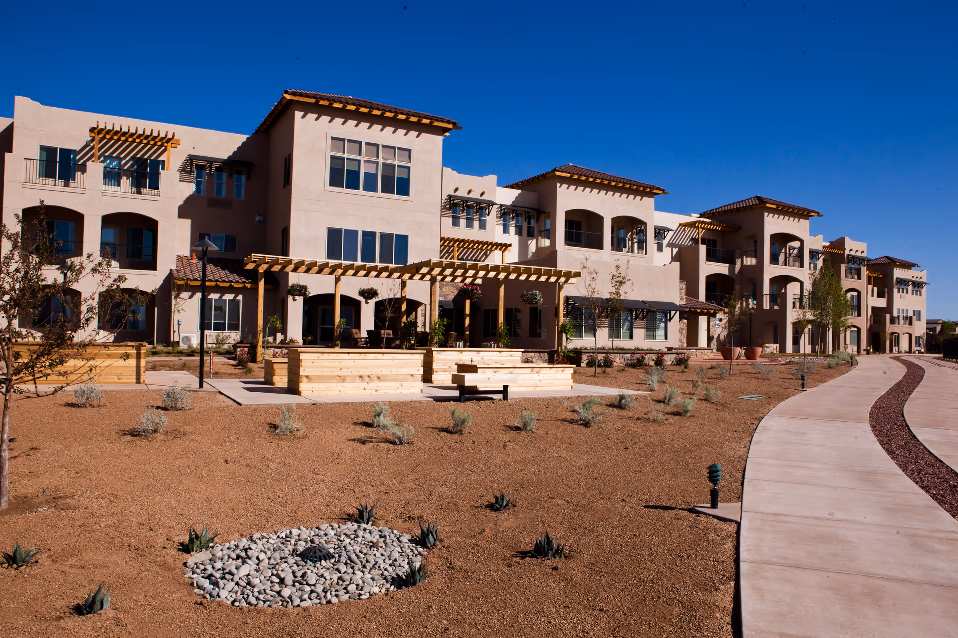 Exterior view of a multi-story retirement living facility with beige stucco walls, wooden pergolas, balconies, and a paved walkway. The landscaping includes desert plants and gravel under a clear blue sky.