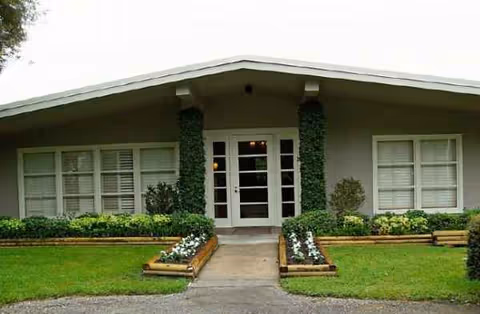 Front entrance of a single-story senior living facility with glass doors, ivy-covered columns, and flower planters.