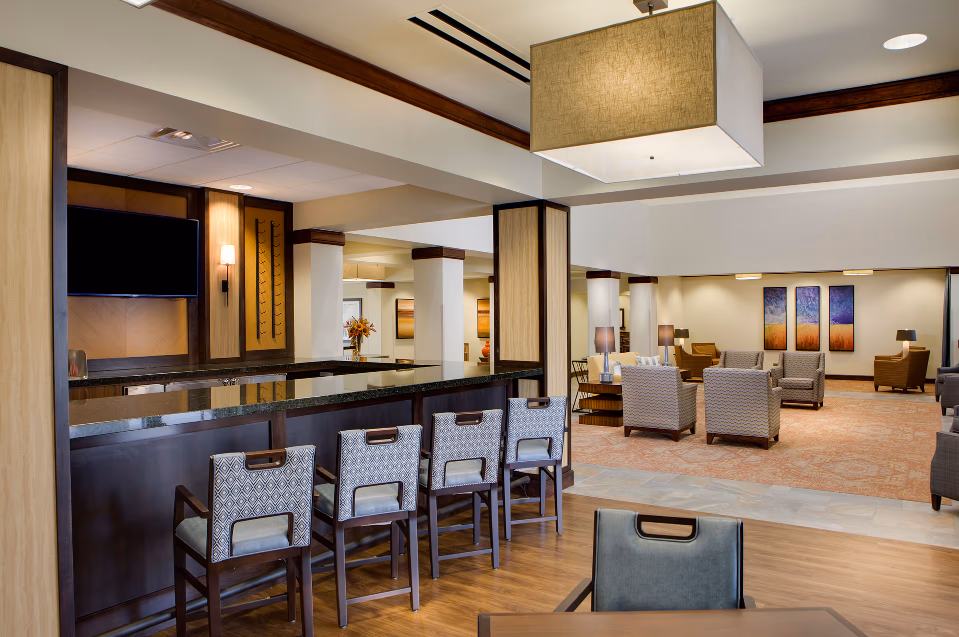 A spacious and well-lit common area in a senior living facility featuring a bar counter with four patterned chairs, a large square ceiling light fixture, and a lounge area with multiple armchairs and side tables. The walls are decorated with colorful abstract paintings, and the flooring transitions from wood near the bar to carpet in the lounge area.