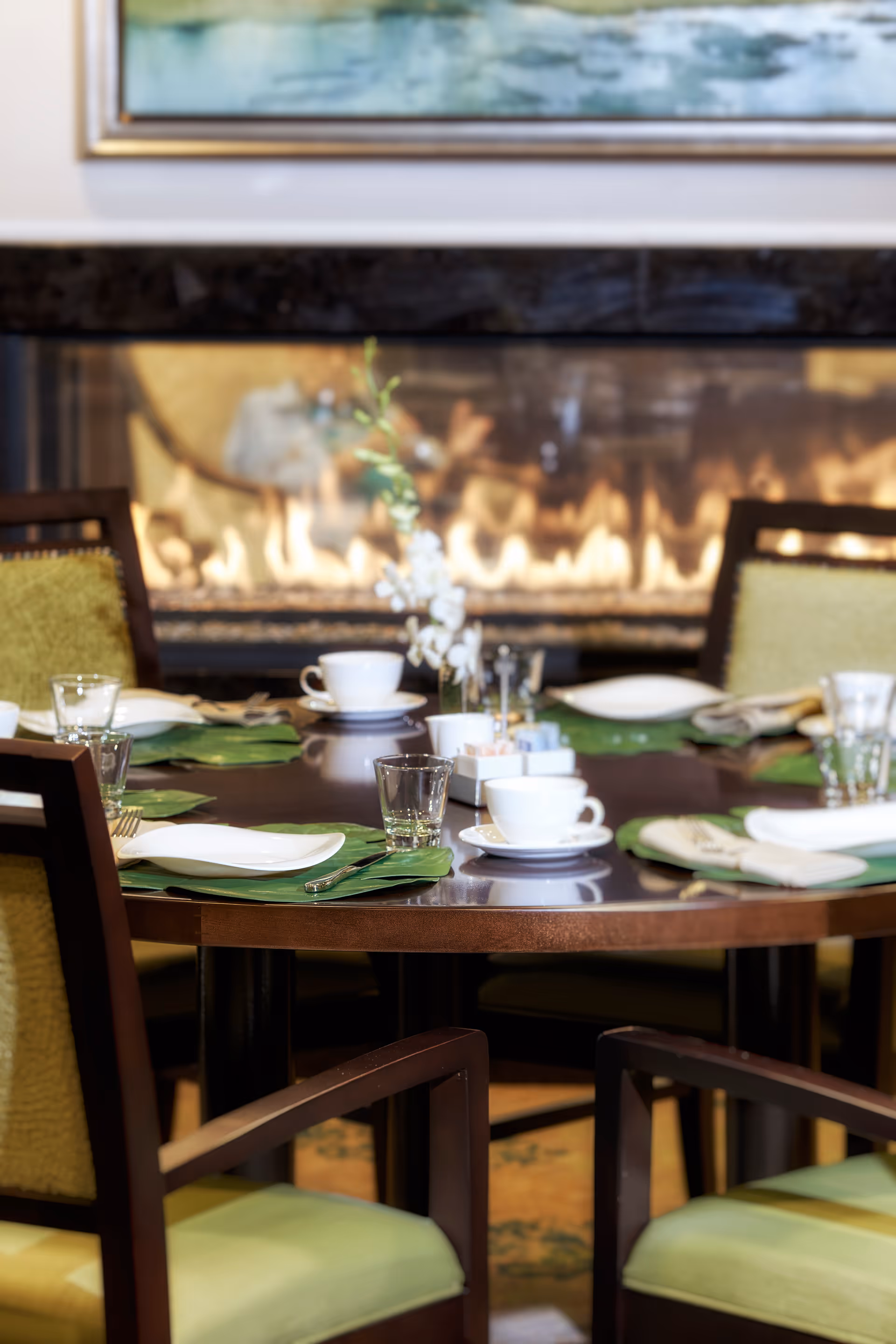 A round dining table set with white cups, glasses, plates, and silverware on green placemats in front of a lit fireplace. The chairs around the table have green cushions and wooden frames.
