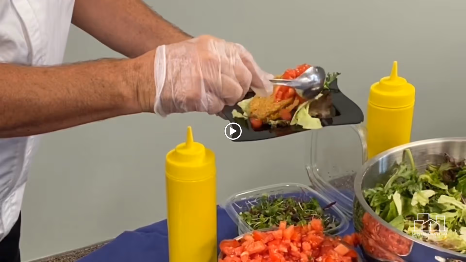 A person wearing a white shirt and disposable gloves is serving a salad with tomatoes and greens onto a black plate. There are two yellow squeeze bottles and containers of chopped vegetables and salad on a table covered with a blue cloth.