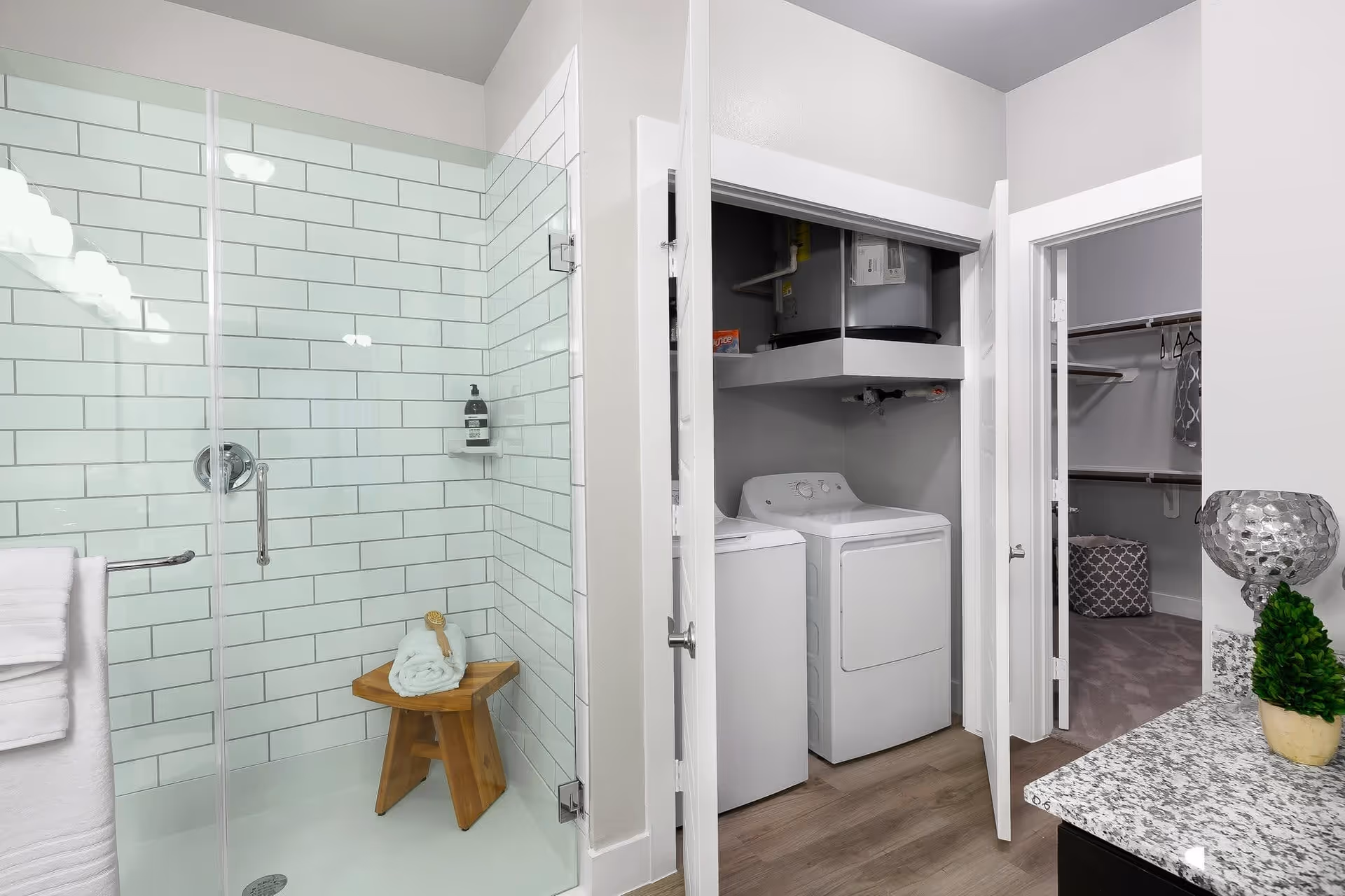 Modern bathroom with a glass-enclosed shower featuring white subway tiles and a wooden stool with a rolled towel and brush. Adjacent to the shower is a closet with a washer, dryer, and water heater. To the right, there is a walk-in closet with hanging rods and a laundry basket. A granite countertop with a decorative plant and lamp is partially visible.