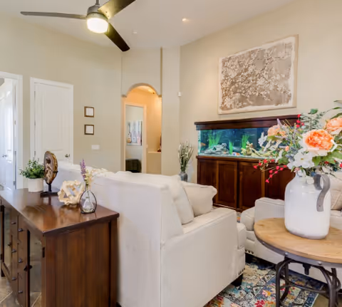 Bright living room with beige sofas, a large aquarium on a wooden cabinet, floral arrangements, and a ceiling fan.