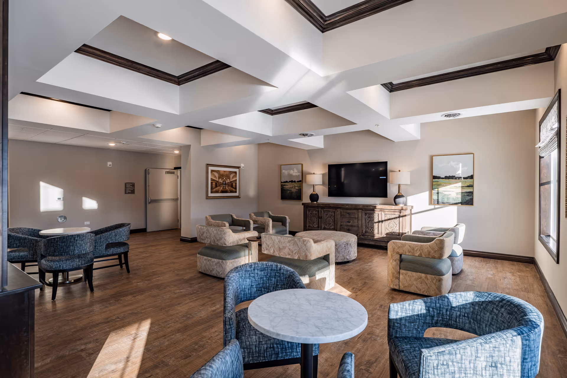 A bright and spacious senior living common area with multiple seating options including blue cushioned chairs around small round tables and beige armchairs with green cushions arranged around a large ottoman. The room features a wooden floor, a coffered ceiling with recessed lighting, a large flat-screen TV mounted on the wall above a wooden cabinet, and framed landscape artwork on the walls. Sunlight streams in through a window on the right side.