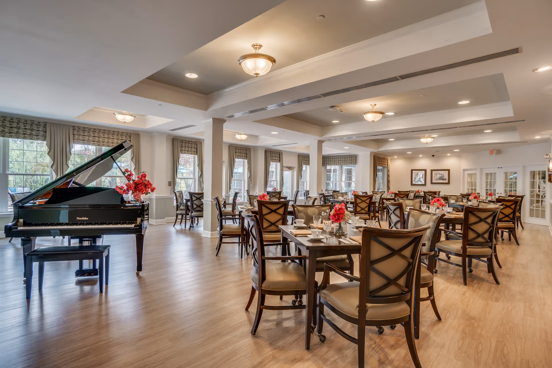 A spacious dining room with multiple wooden tables and chairs arranged neatly. Each table is set with glassware, cups, and floral centerpieces. Large windows with curtains allow natural light to fill the room. A black grand piano with a bouquet of red flowers on top is positioned near the windows. The ceiling features recessed lighting and decorative light fixtures.
