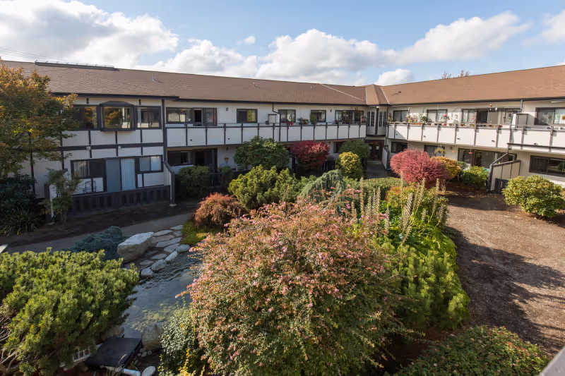 View of a senior living facility courtyard with a variety of green shrubs, bushes, and trees. The two-story building surrounds the courtyard, featuring balconies and windows overlooking the garden area under a partly cloudy sky.