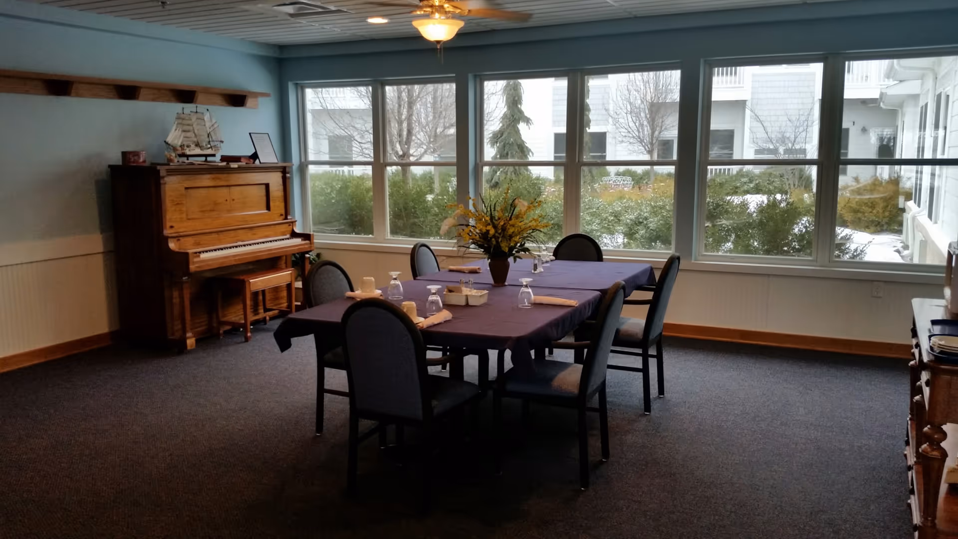 A dining room with a rectangular table covered with a purple tablecloth, set with six chairs, napkins, glasses, and cups. A vase with yellow flowers is in the center of the table. Large windows show an outdoor view with bushes and trees. A wooden piano with a model ship on top is against the left wall, and a ceiling fan with a light hangs above.