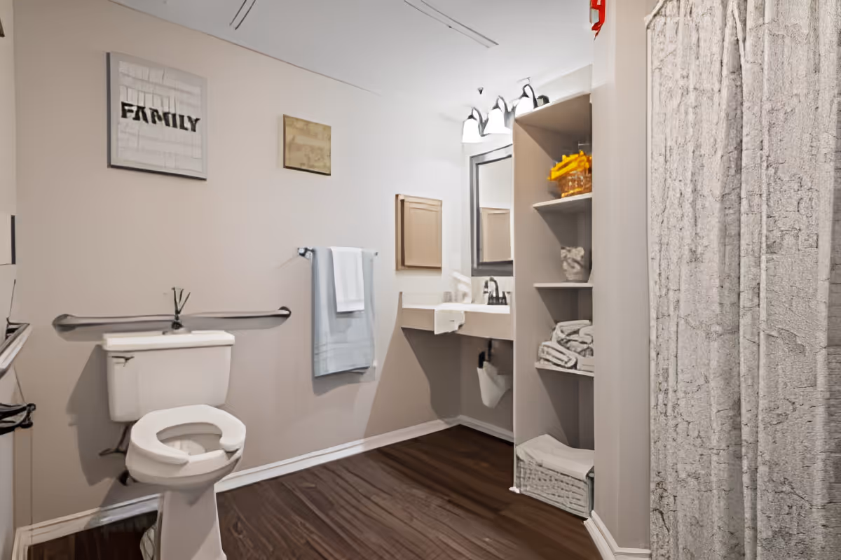 A clean and accessible bathroom with a toilet featuring a raised seat and grab bars, a sink with a mirror above it, shelves stocked with towels and toiletries, and a shower with a textured curtain. The walls are decorated with framed artwork, including one with the word 'FAMILY'.