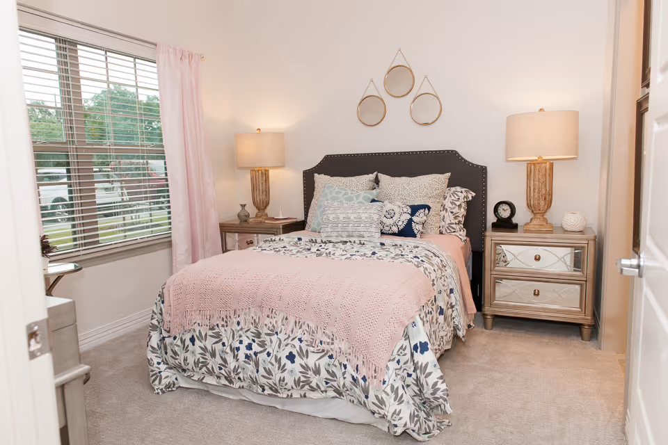 Cozy bedroom with a floral-patterned bed topped by a pink throw, flanked by two nightstands with lamps and a window on the left.