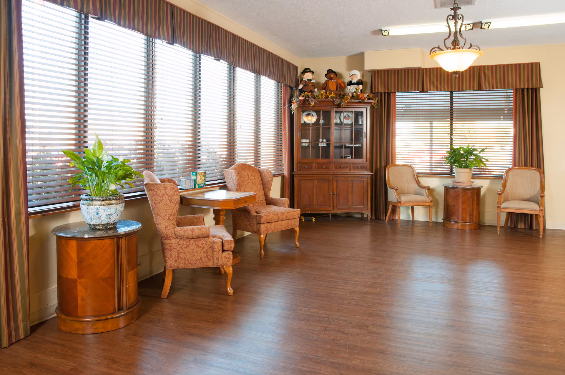 A bright and spacious common area with large windows covered by blinds and striped valances. The room features wooden flooring, two upholstered armchairs with a small wooden table between them, a wooden cabinet with decorative items on top, and two additional chairs with a small round table holding a potted plant. A ceiling light fixture hangs from above.