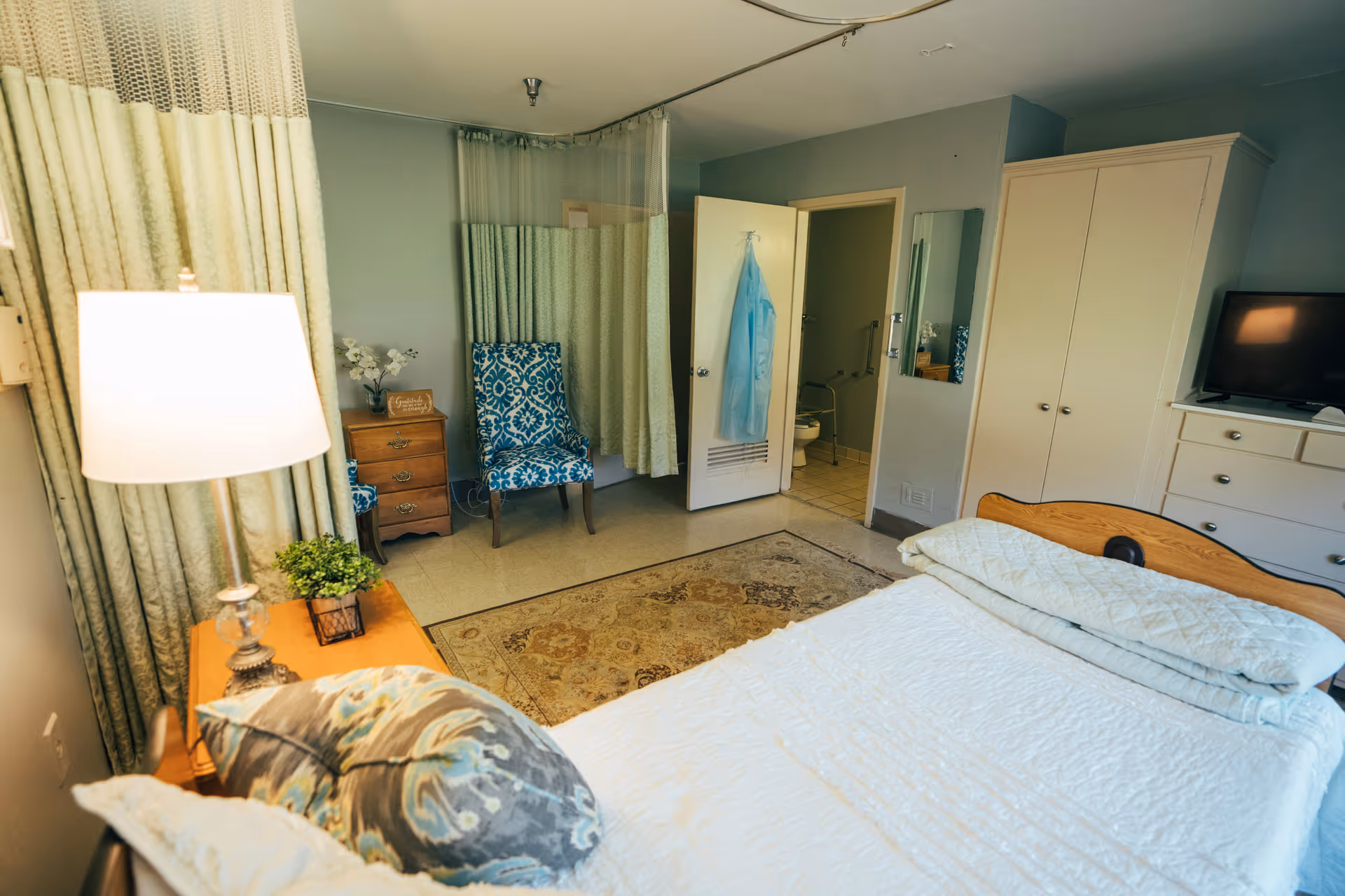 A cozy bedroom in a senior living facility with a bed covered in white bedding, a bedside table with a lamp and a small plant, a patterned blue chair, a wooden dresser with a TV on top, a wardrobe, and an open door leading to a bathroom with grab bars and a blue gown hanging on the door.