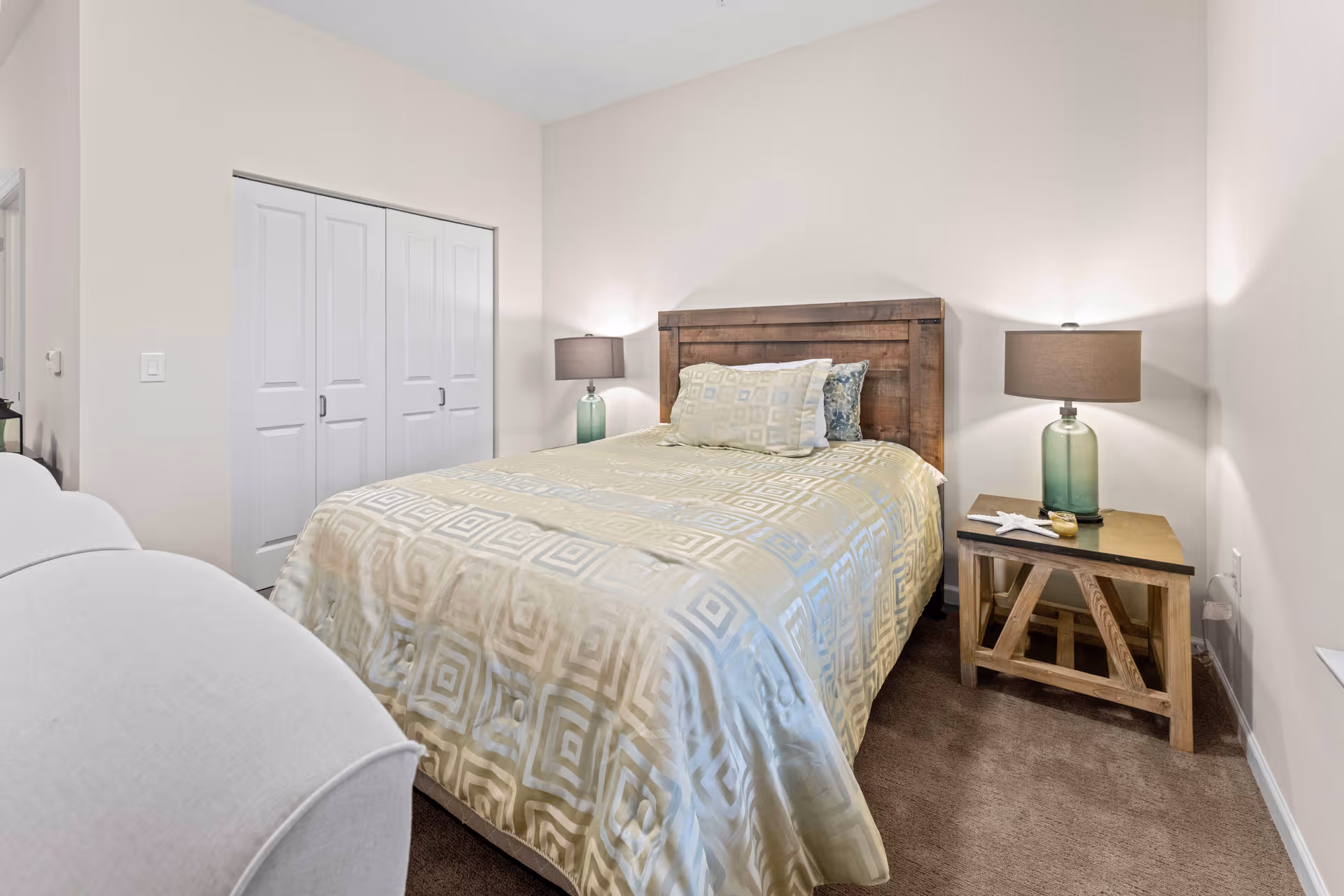 A bedroom with a wooden headboard bed covered with a beige and white patterned comforter and pillows. There are two bedside tables with green glass lamps and a starfish decoration on the right table. A white closet with sliding doors is visible on the left wall, and a white armchair is partially visible in the foreground.